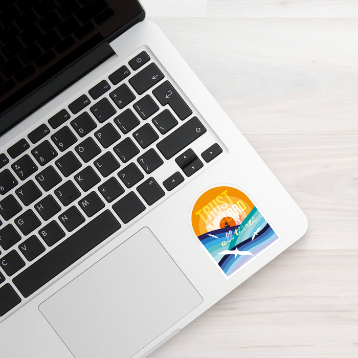 A round sticker with a retro sunset beach graphic and the text 'TRUSTED by Ocean' applied to the lower right corner of a silver laptop palm rest, placed on a light wood surface.