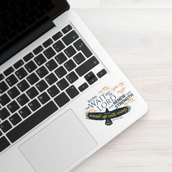 A close-up of a silver laptop on a light wood surface, with a decorative sticker applied to the palm rest. The sticker features a raven and the text 'WAIT FOR THE LORD AND RENEW YOUR STRENGTH; heart of trust with Him' in black, white, and orange.