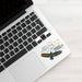 A close-up of a silver laptop on a light wood surface, with a decorative sticker applied to the palm rest. The sticker features a raven and the text 'WAIT FOR THE LORD AND RENEW YOUR STRENGTH; heart of trust with Him' in black, white, and orange.