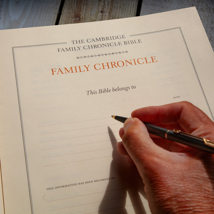 An open book showing the Family Chronicle page from The Cambridge Family Chronicle Bible, with a hand holding a black and gold pen writing on the lined page, set on a dark wooden surface.
