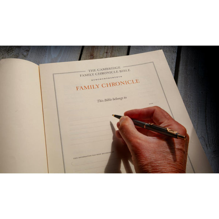 An open copy of The Cambridge Family Chronicle Bible on a dark wooden surface, with a hand holding a black and gold pen writing on the page.
