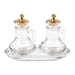Two clear glass prayer water bottles with gold cross lids, set on a matching clear glass tray against a white background.