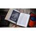 An open Holy Bible lies on a rustic wooden surface, flanked by a closed black Holy Bible on the left and a closed red/orange Family Bible on the right. The open Bible shows a black-and-white illustration on the left page and a text page on the right.