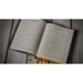 An open hardcover journal or calendar laid on a rustic wooden surface, showing printed calendar pages with sections for monthly and yearly summaries. The book has a dark patterned cover and ribbon bookmarks.