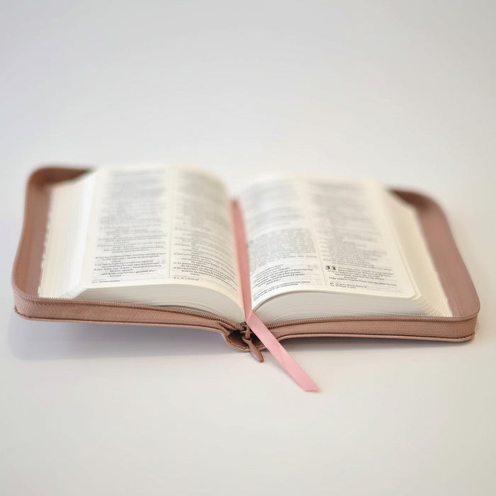An open zippered Bible or book cover in a muted pinkish-brown leather-like material, with white pages spread open and a pink ribbon bookmark hanging from the center, photographed on a light neutral background.