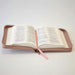 An open zippered Bible or book cover in a muted pinkish-brown leather-like material, with white pages spread open and a pink ribbon bookmark hanging from the center, photographed on a light neutral background.